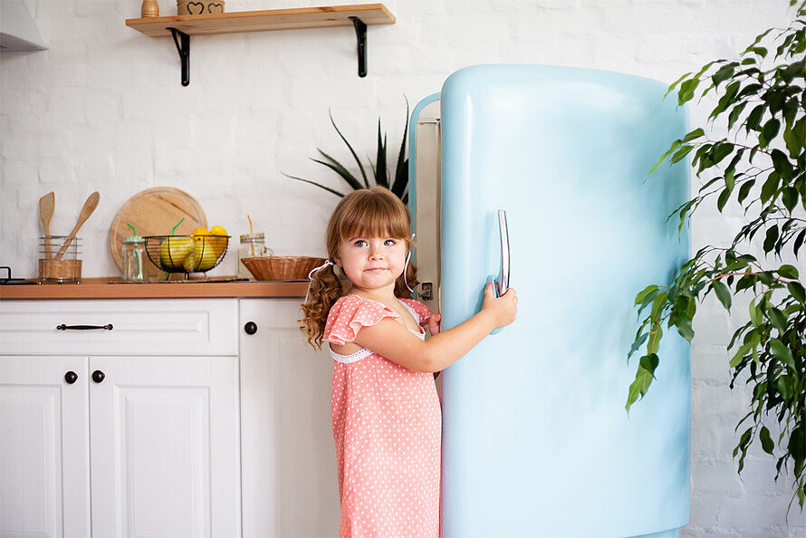 Ein rothaariges Mädchen steht an einer geöffneten Kühlschranktür und blick in die Kamera.