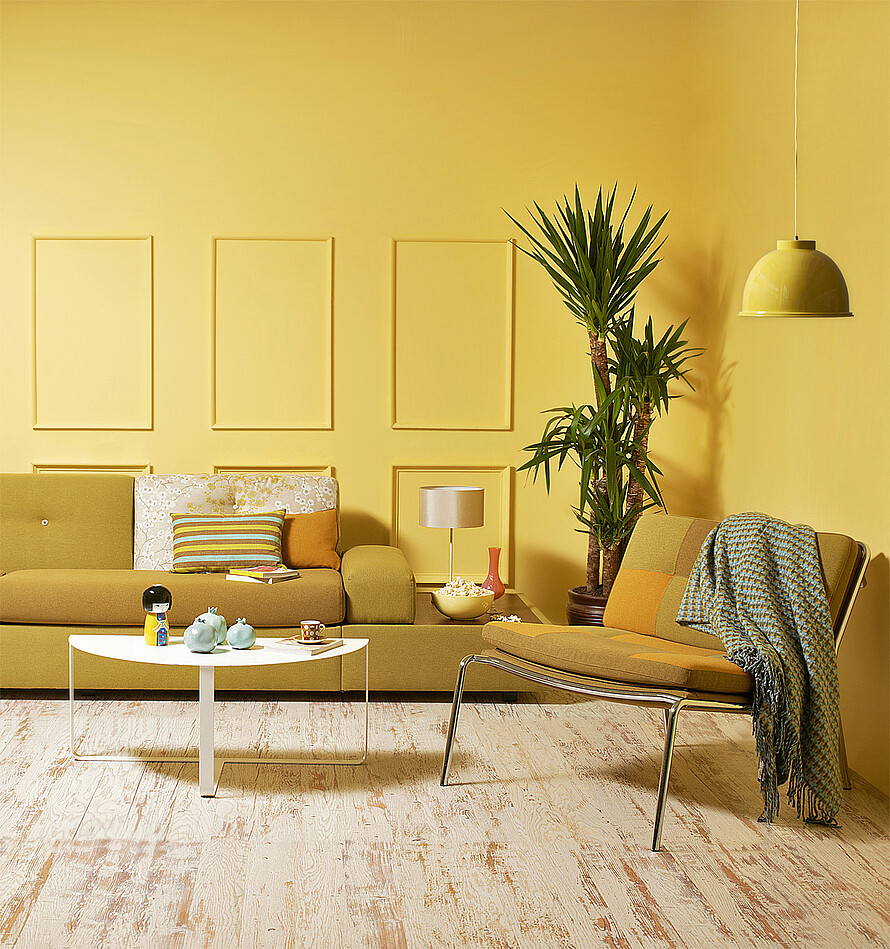 Ein Wohnzimmer mit senfgelber Couch, Couchtisch, Sessel und diversen Accessoires. Im Fokus steht die gelbe Wandfarbe, die sich über die gesamte Wandfläche verteilt.