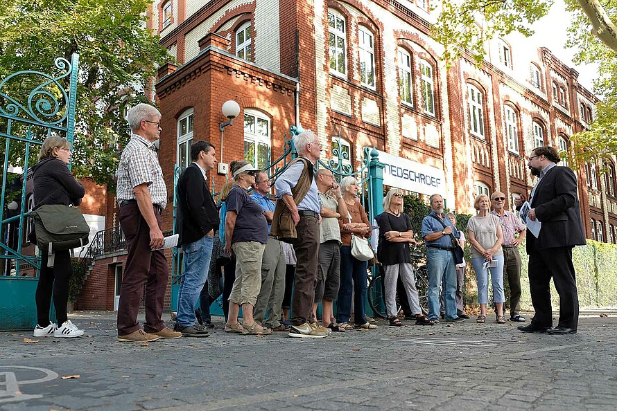 Eine Gruppe von Menschen steht auf einem gepflasterten Weg vor einem rot-weiß gemauerten Gebäude. Eine Person steht vor der Gruppe und erzählt etwas.