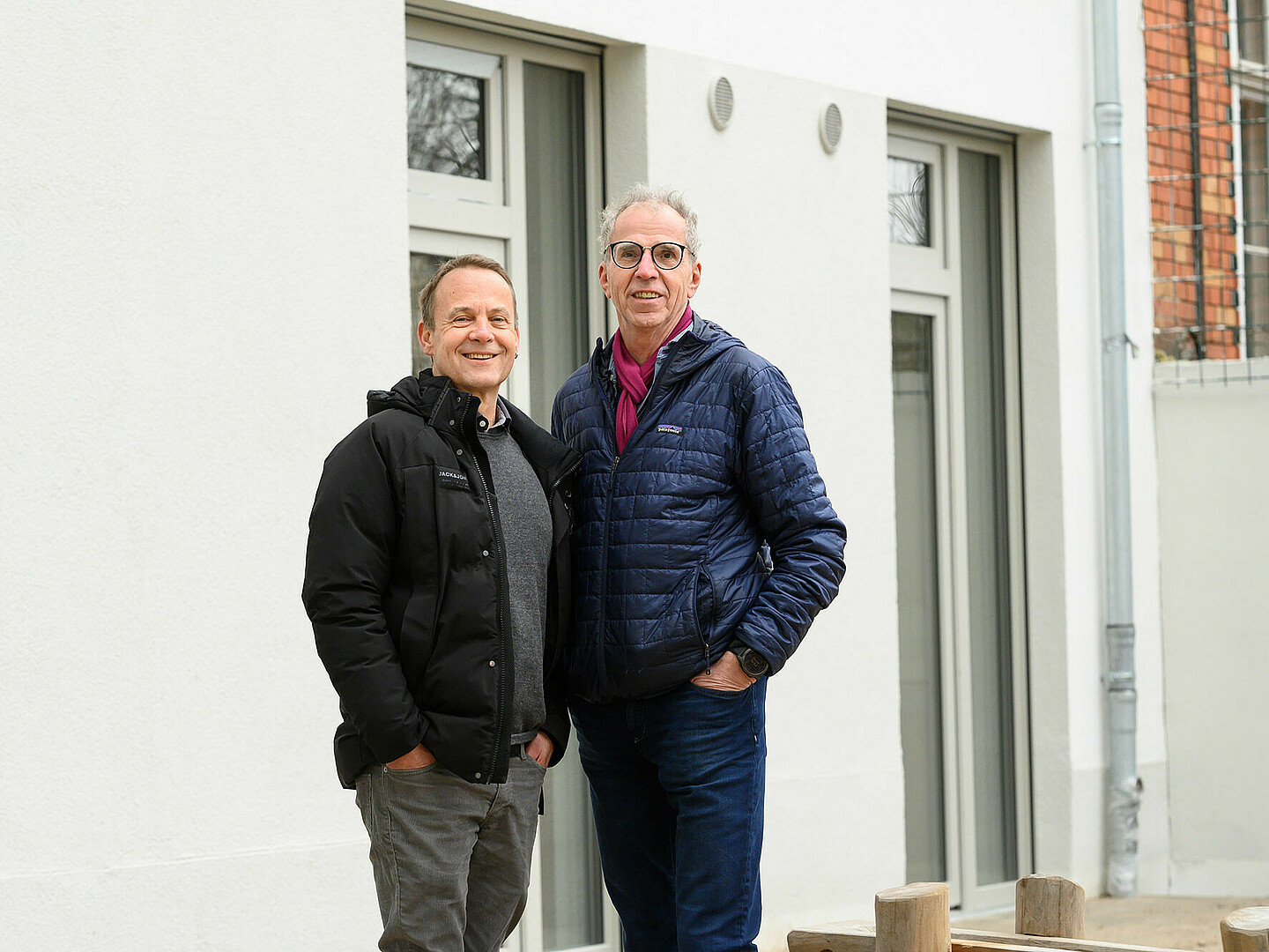 Zwei Männer stehen vor einer weißen Gebäudewand und lachen in die Kamera.