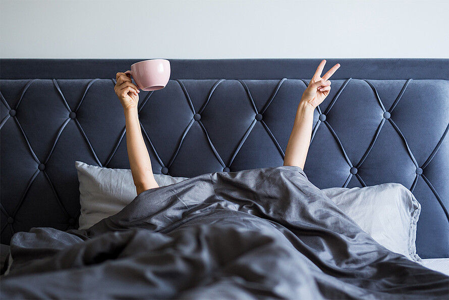 Ein großes, graues Bett im Anschnitt, eine Person liegt unter der Decke, nur zwei Arme sind zu sehen. In ihrer rechten Hand hält die Person eine große Tasse.