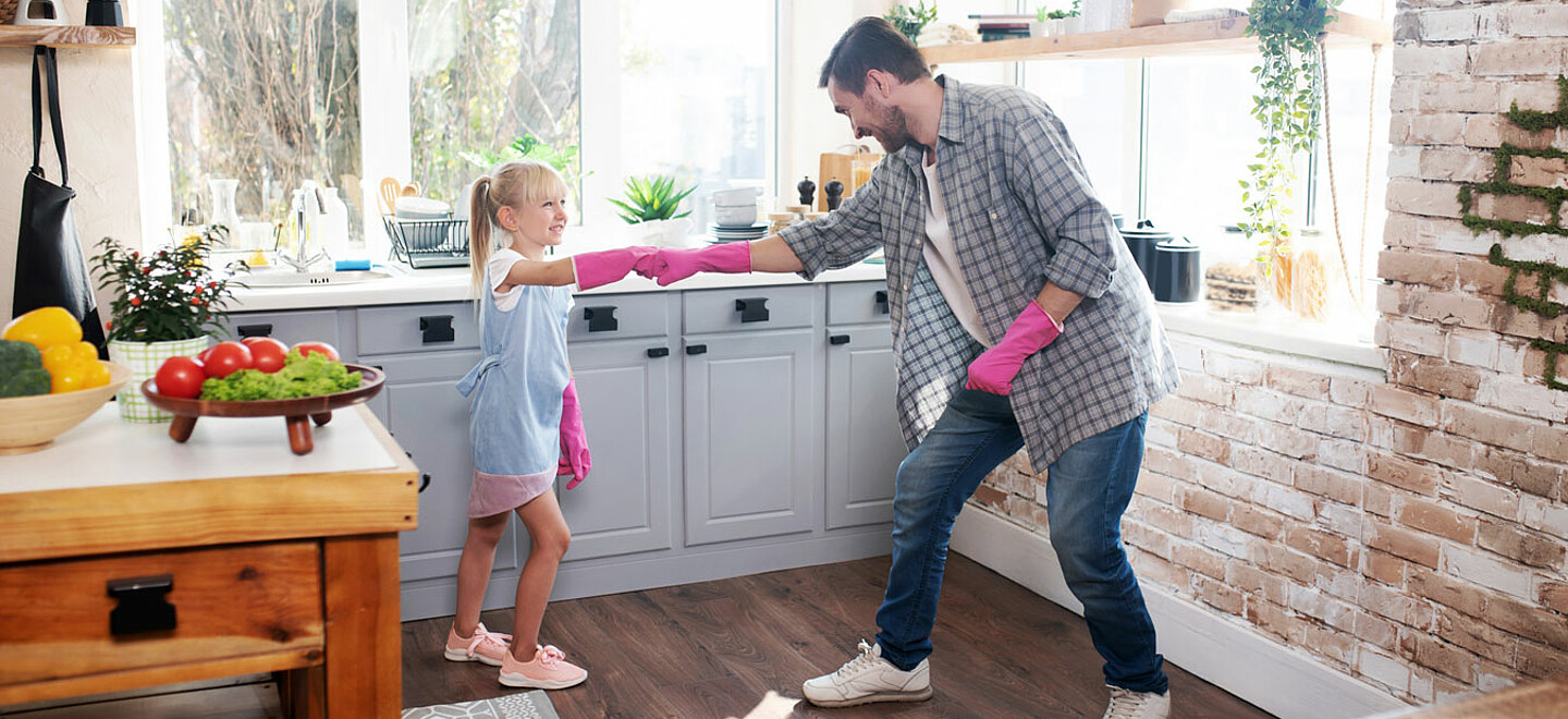Das Foto zeigt Vater und Tochter mit pinken Gummihandschuhen in einer Küche. Beide lachen und machen einen Faustgruß.
