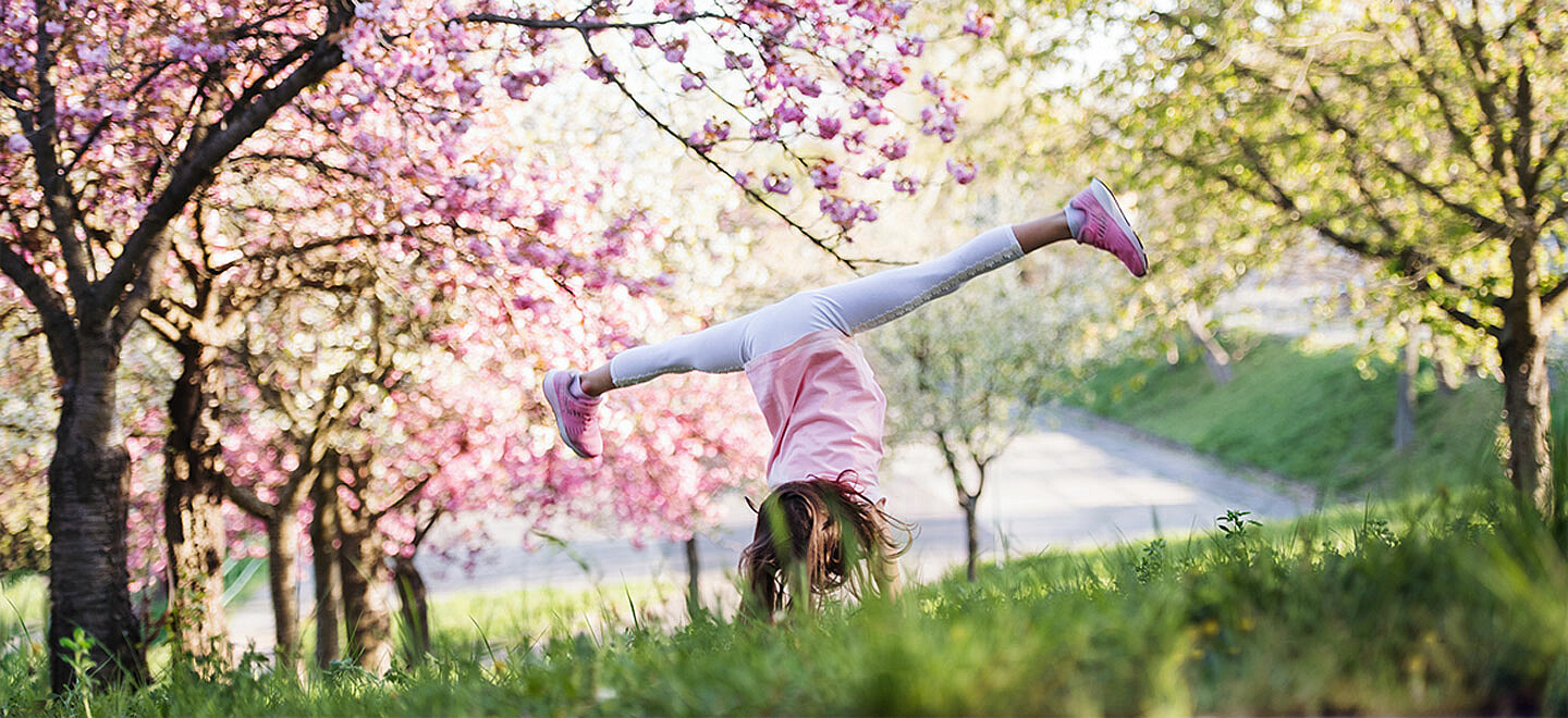 Mädchen macht einen Radschlag auf einer grünen Wiese. Im Hintergrund blühen die Bäume im Frühling.