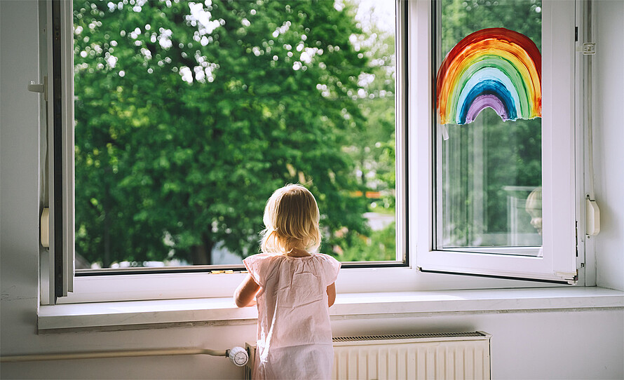 Ein Kind steht vor einem geöffneten Fenster. Auf die Fensterscheibe wurde ein Regenbogen gemalt.