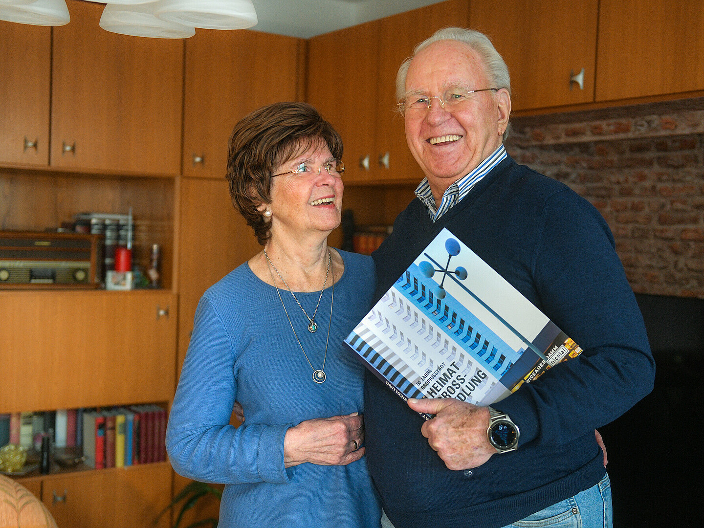 Bärbel und Udo Schulte in ihrem Wohnzimmer, Udo Schulte hält einen Bildband anlässlich des 50. Jubiläums der Gropiusstadt in der Hand.