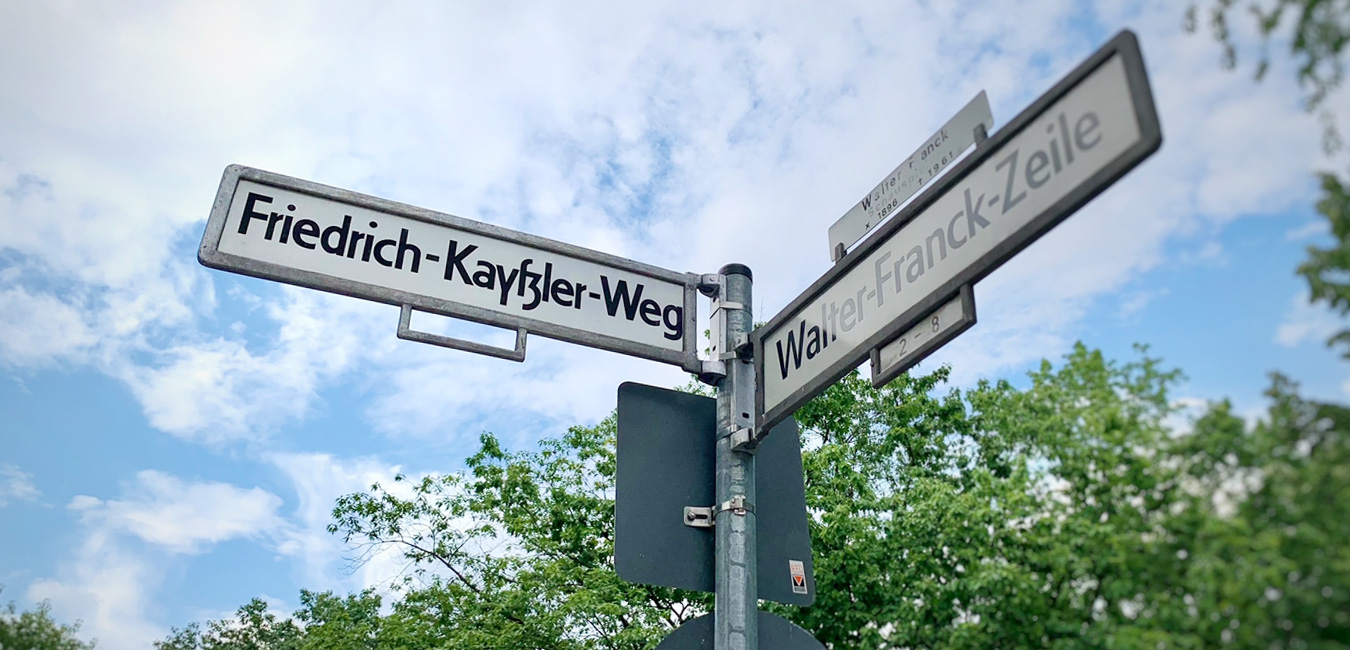 Ein Straßenschild mit der Aufschrift „Friedrich-Kayßler-Weg“. Daneben ein Straßenschild mit der Aufschrift „Walter-Frank-Zeile“. Im Hintergrund blauer Himmel, ein paar Wolken und Baumkronen.