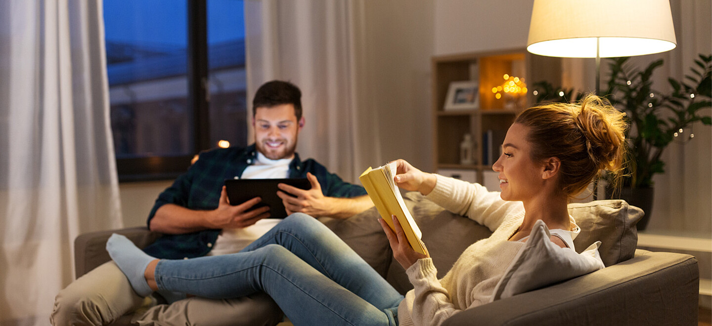 Eine Frau und ein Mann sitzen auf der Couch. Der Mann schaut auf sein Tablet und die Frau liest ein Buch. Im HIntergrund steht eine Lampe.