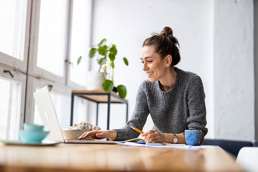 Eine lächelnde Frau sitzt an einem Schreibtisch, der am Fenster steht. Auf dem Schreibtisch steht ein Laptop, sowie eine Tasse und weitere Arbeitsgegenstände. Im Hintergrund ist eine Pflanze zu sehen. Der Raum ist sehr hell.