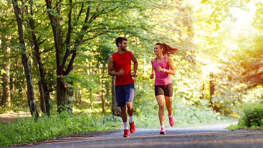 Zwei Personen joggen durch einen Park, der Weg ist gesäumt von vielen großen Bäumen.