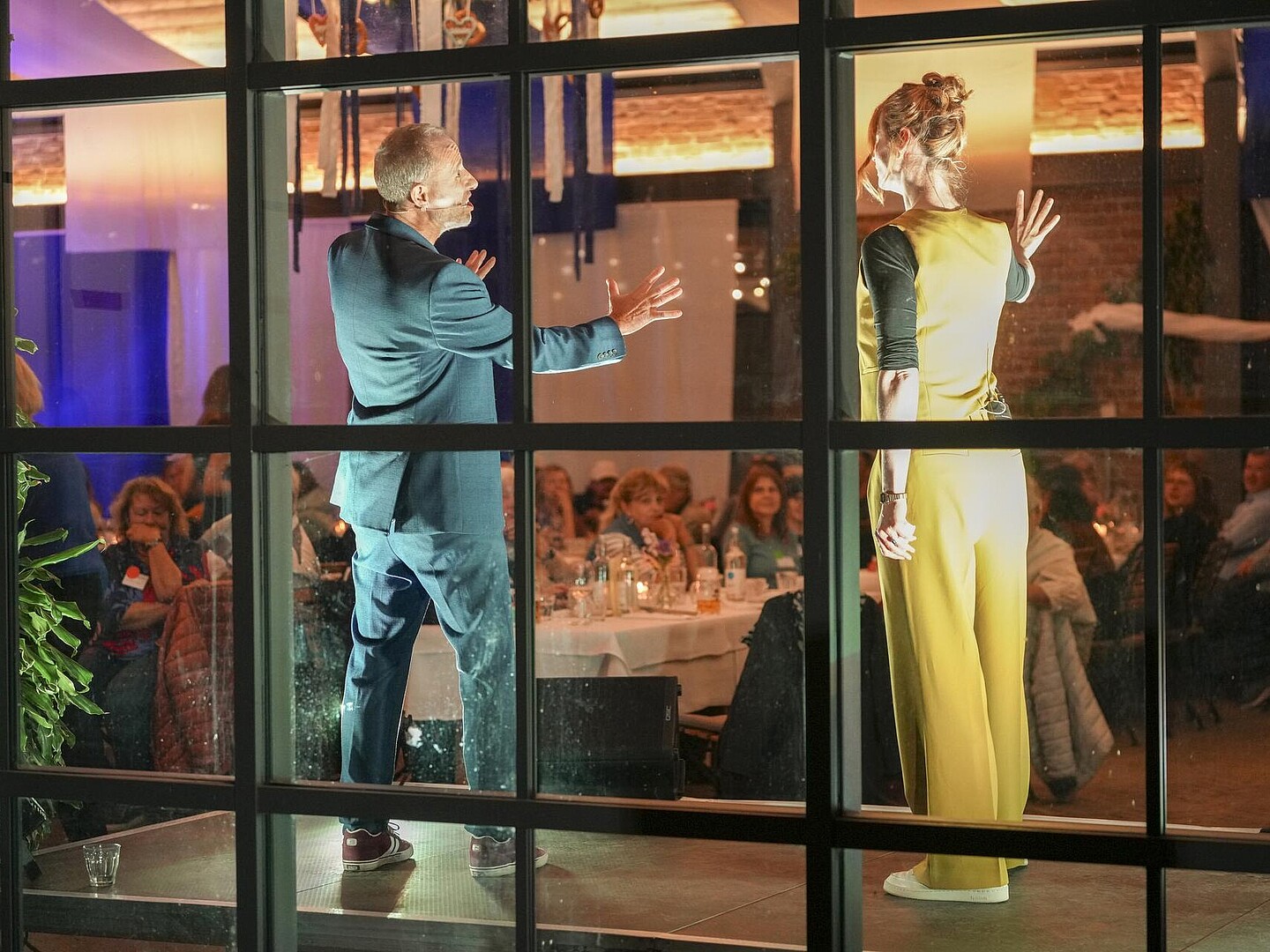 Blick von außen durch die Fensterscheibe nach innen in das Neue Café, während das Theater auf der Bühne spielt.