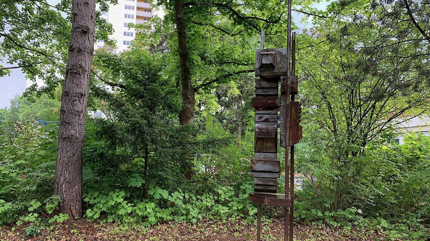 Eine abstrakte Skulptur aus teilweise rostigem Stahl die vor grünen Bäumen und Sträuchern steht.