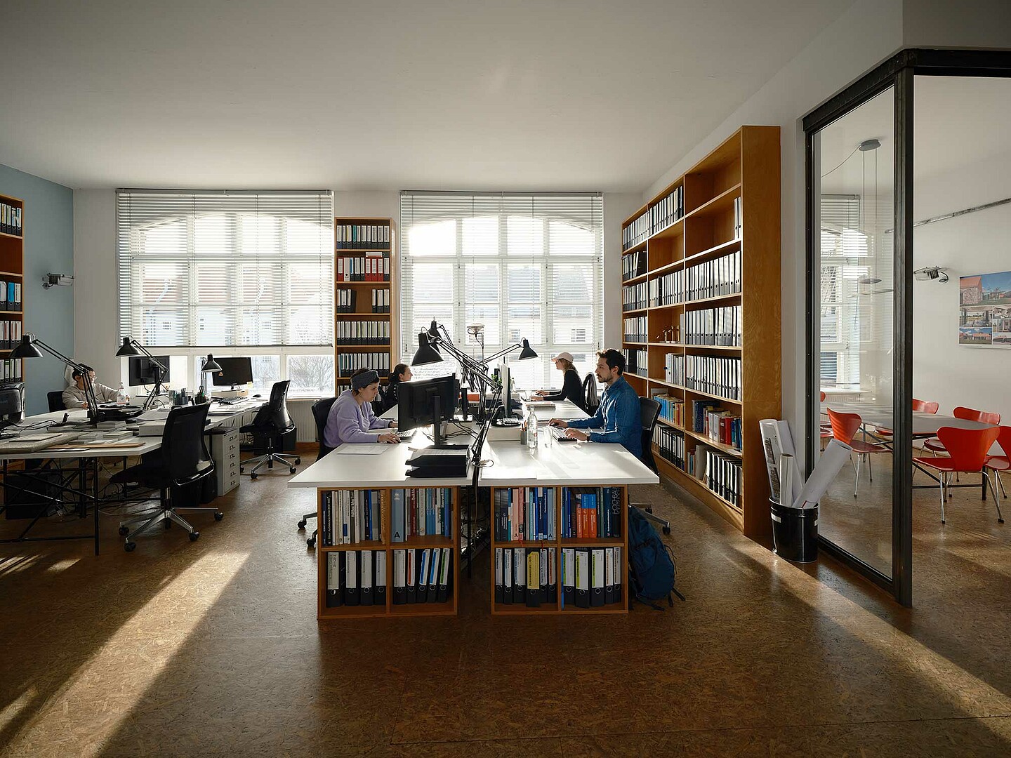 Vier Menschen arbeiten in einem geräumigen Architekturbüro. Sie sitzen am Computer und sind vertieft in ihre Arbeit. An den Wänden stehen hohe Regale, die mit Büchern und Ordnern gefüllt sind.  Durch die großen Fenster fällt viel Licht.