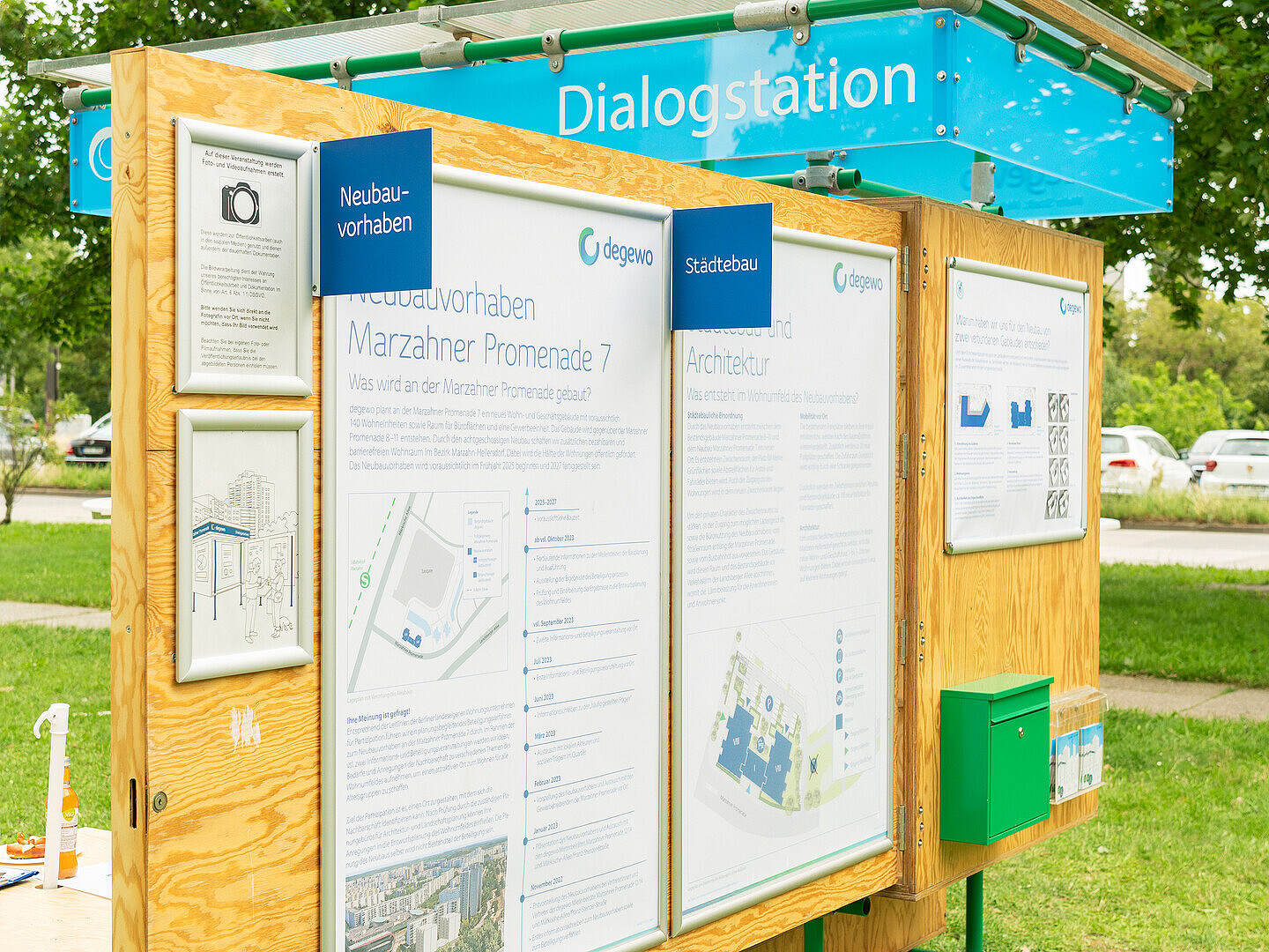 Die degewo-Dialogstation ist im aufgeklappten Zustand. An einer Holzwand hängen mehrere große Plakate, daneben ein Briefkasten. Auf der Rückseite ein kleiner Tisch mit Hockern.