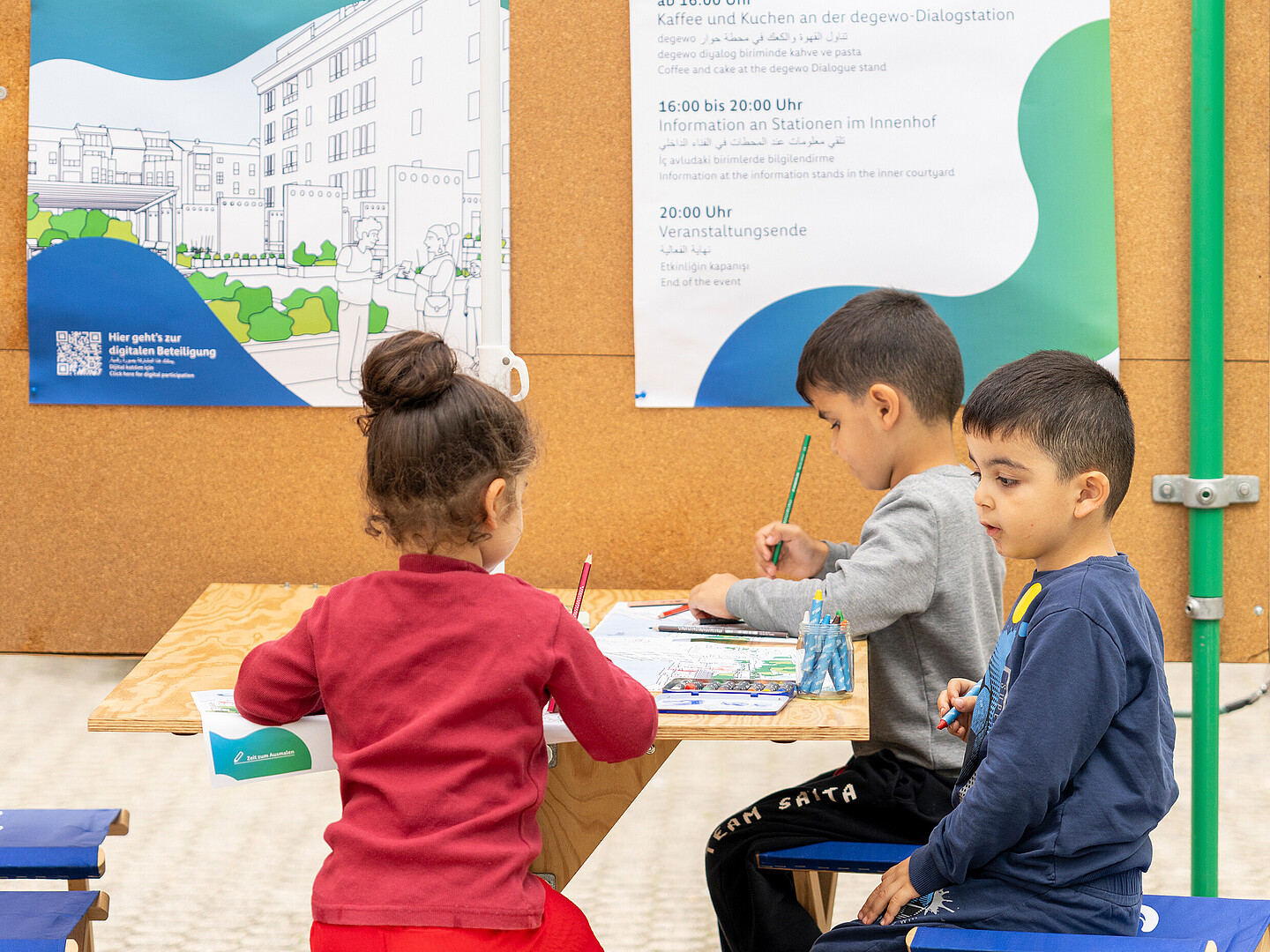 Drei Kinder sitzen an einem Tisch und malen. Hinter dem Tisch eine große Korkwand mit zwei degewo-Plakaten. Über den Kindern ein Sonnenschirm mit degewo-Logo.