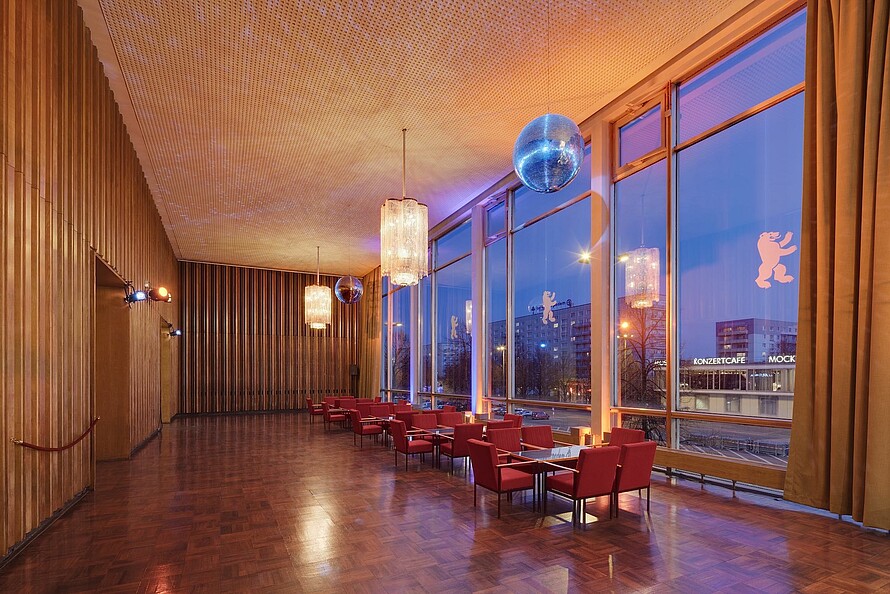 Ein Foyer vor großen Fenstern mit roten Stühlen und Tischen im Stil der sechziger Jahre, schimmerndem Parkett und holzvertäfelten Wänden. Von der Decke baumeln prunkvolle Lampen und eine Diskokugel.