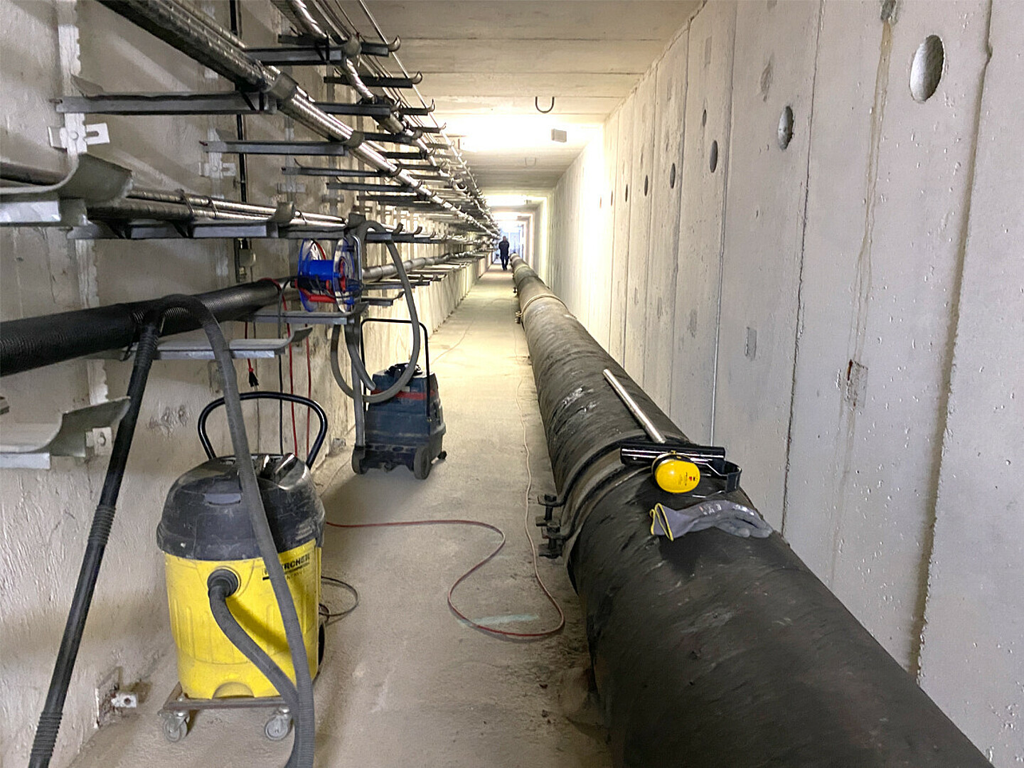 Im Sammelkanal in Marzahn steht ein Staubsauger, am Boden entlang verläuft ein großes Rohr, an der Wand verschiedene Kabel auf Kabelpfannen.