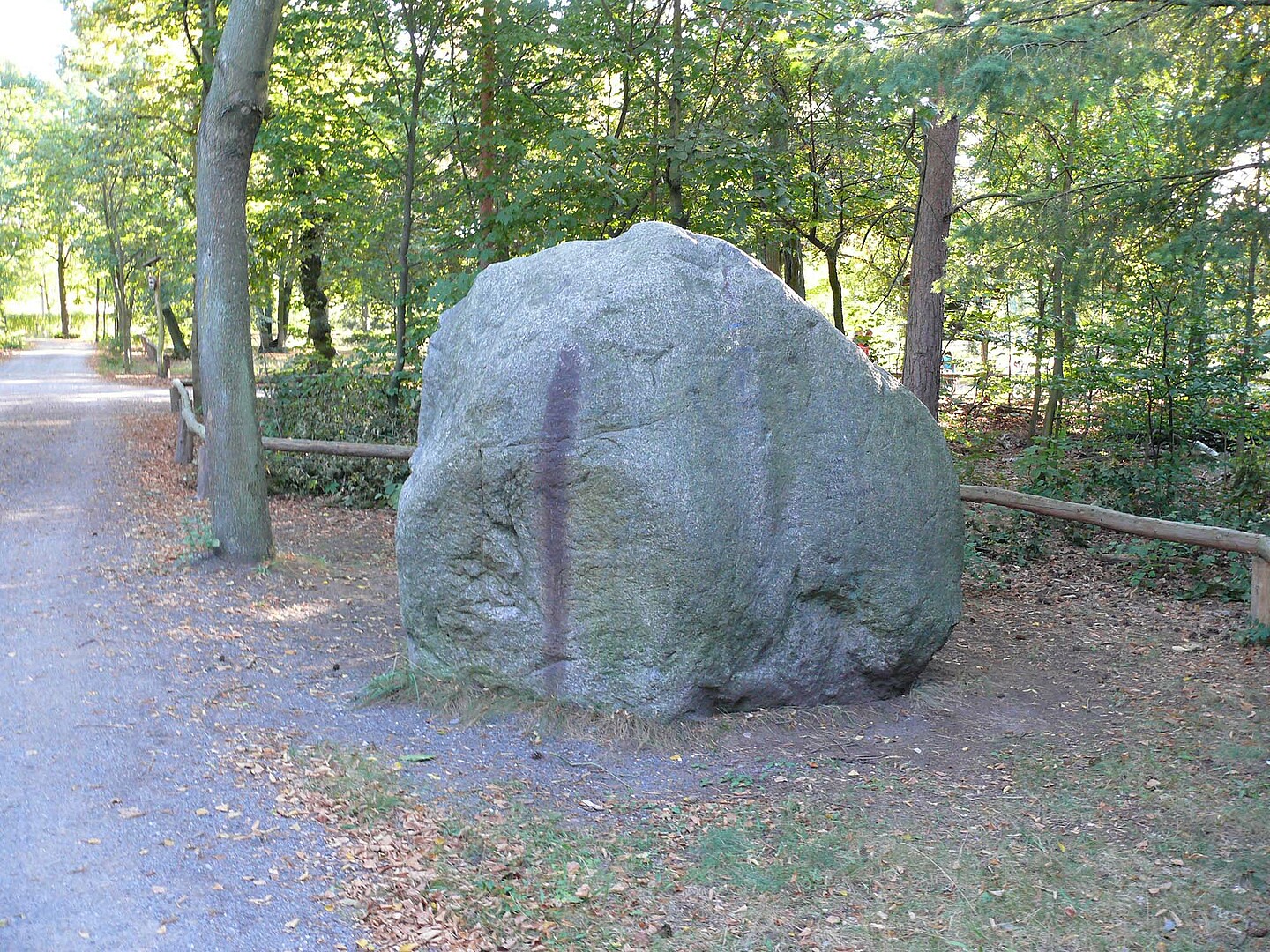 Ein grauer großer Stein auf einer Wiese neben einem Waldweg.