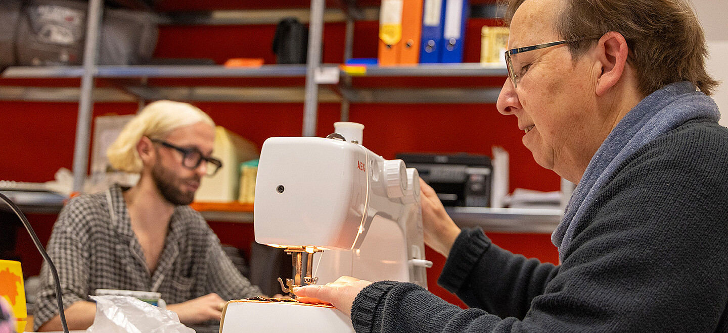 Eine Mitarbeiterin sitzt an der Nähmaschine, im Hintergrund arbeitet ein Mann am Computer.