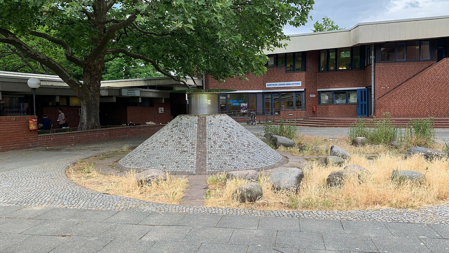 Eine kegelförmige Skulptur, die mit hellen Ziegeln verkleidet ist. Auf der Spitze ein zylinderförmiger Aufbau aus einem anderen Material. Im Hintergrund ein Gebäude und ein Baum.