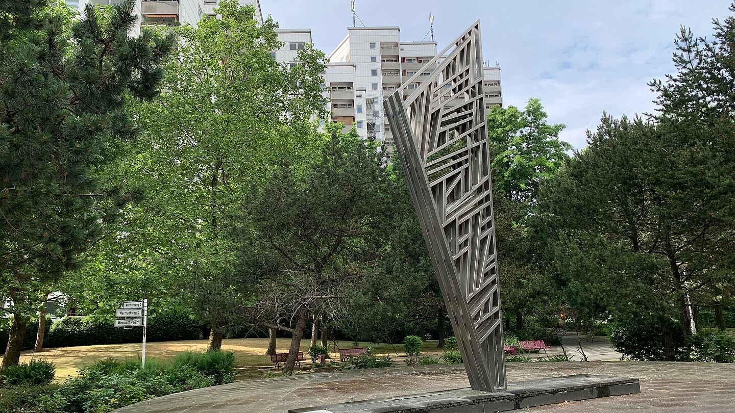 Eine abstrakte Stahlskulptur mit vielen unterschiedlich angeordneten Streben auf einem runden, flachen Podest, das begehbar ist. Im Hintergrund Bäume und hohe Wohnhäuser.