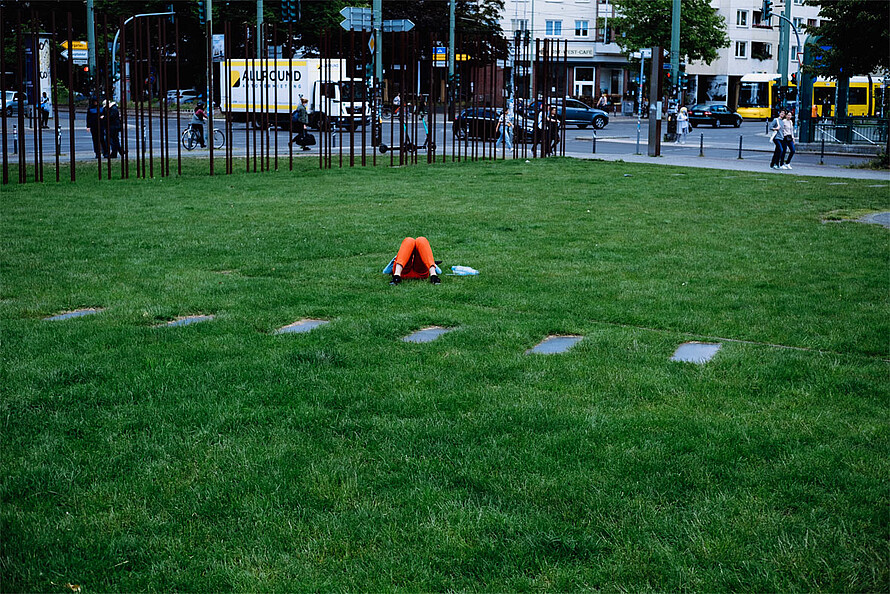 Das Foto zeigt eine grüne Wiese, in deren Mitte eine Person entspannt liegt. Das Fleckchen Grün liegt direkt an einer Straßenkreuzung an der Bernauer Straße.