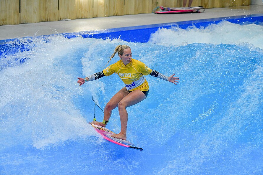 Eine junge Frau surft im Wasserbecken einer Indoor-Halle auf einem Surfbrett.