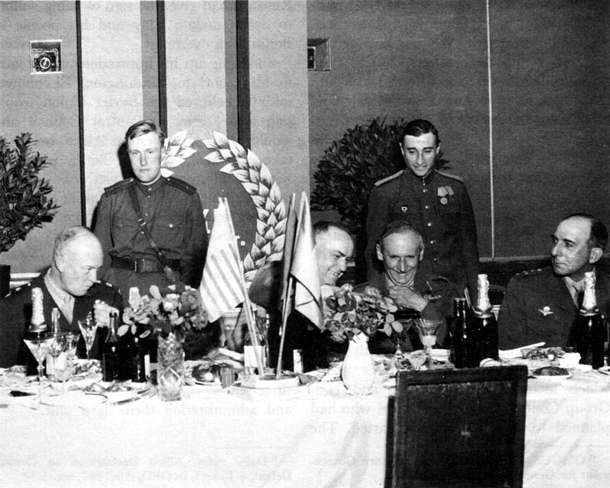 Scharz-weiß Fotografie von sechs Personen. Vier sitzen an einem gedeckten Tisch, zwei Stehen dahinter.