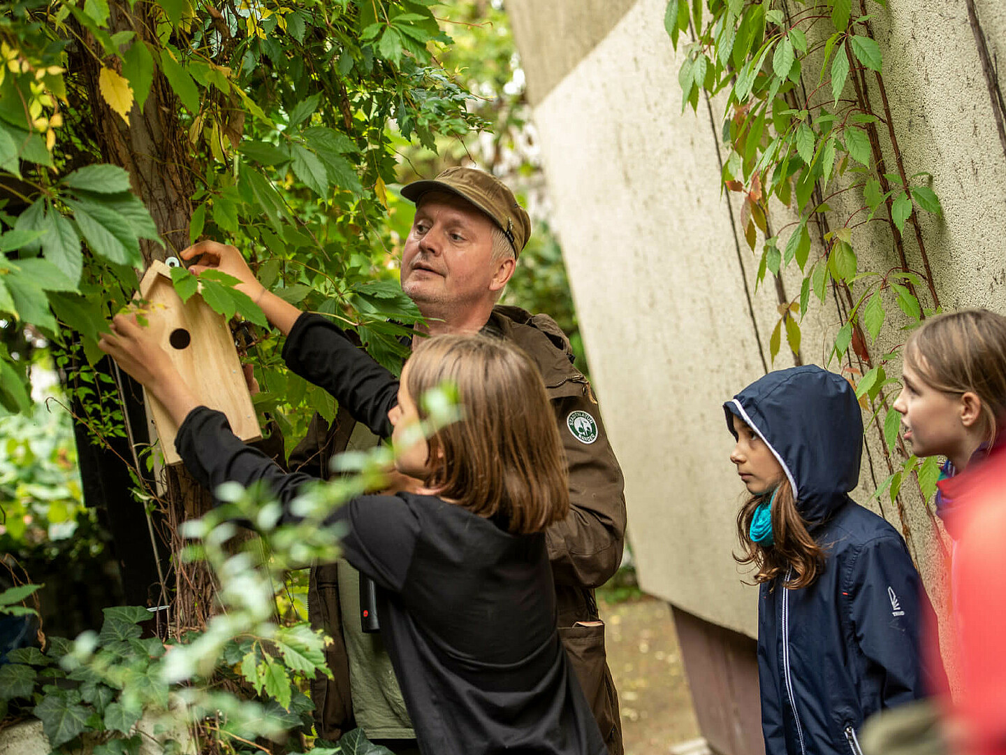 Mann hilft Kindern ein Vogelhaus an einen Baum anzubringen.