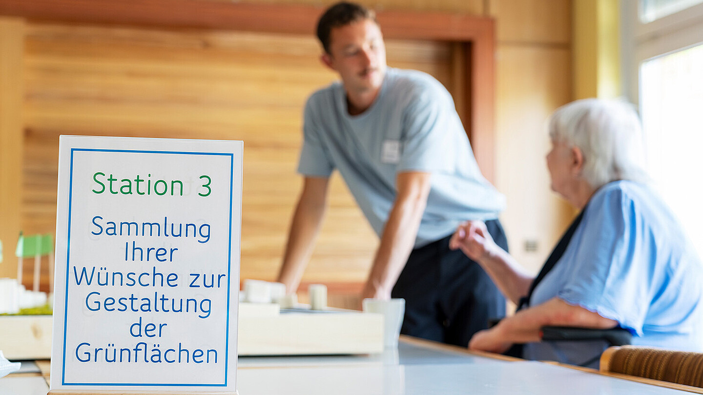 Eine ältere Frau sitzt an einem Tisch in einem Raum und lässt sich von einem Mitarbeiter von degewo beraten. Auf einem Tischaufsteller steht Station 3, Sammlung Ihrer Wünsche zur Gestaltung der Grünflächen.