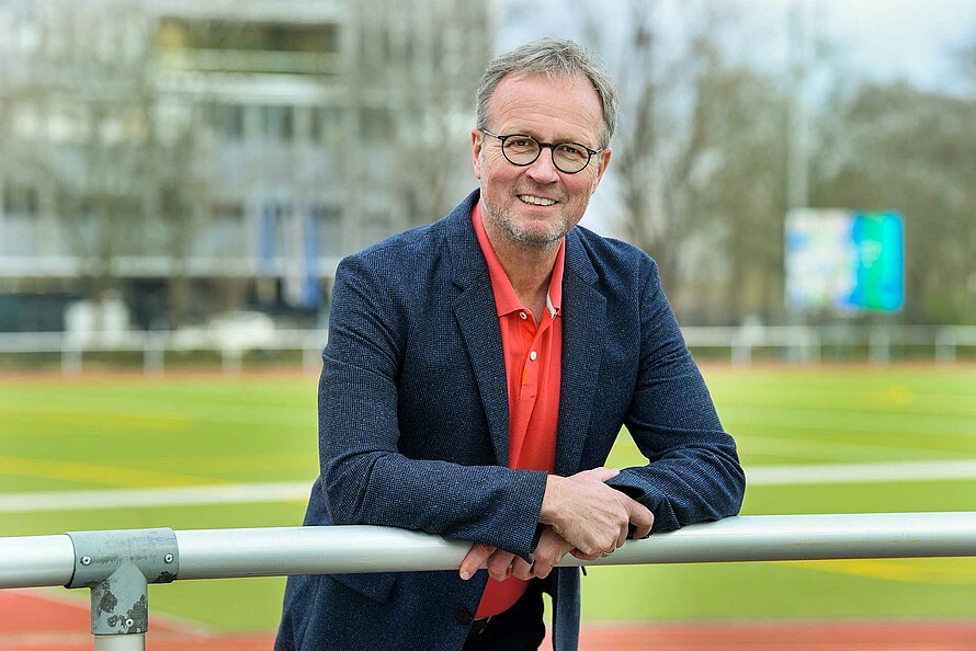 Martin Seeber hat seine Arme auf ein Geländer gelegt. Er lächelt in die Kamera. Im Hintergrund ist verschwommen der Rasen eines Fußballplatzes zu sehen.