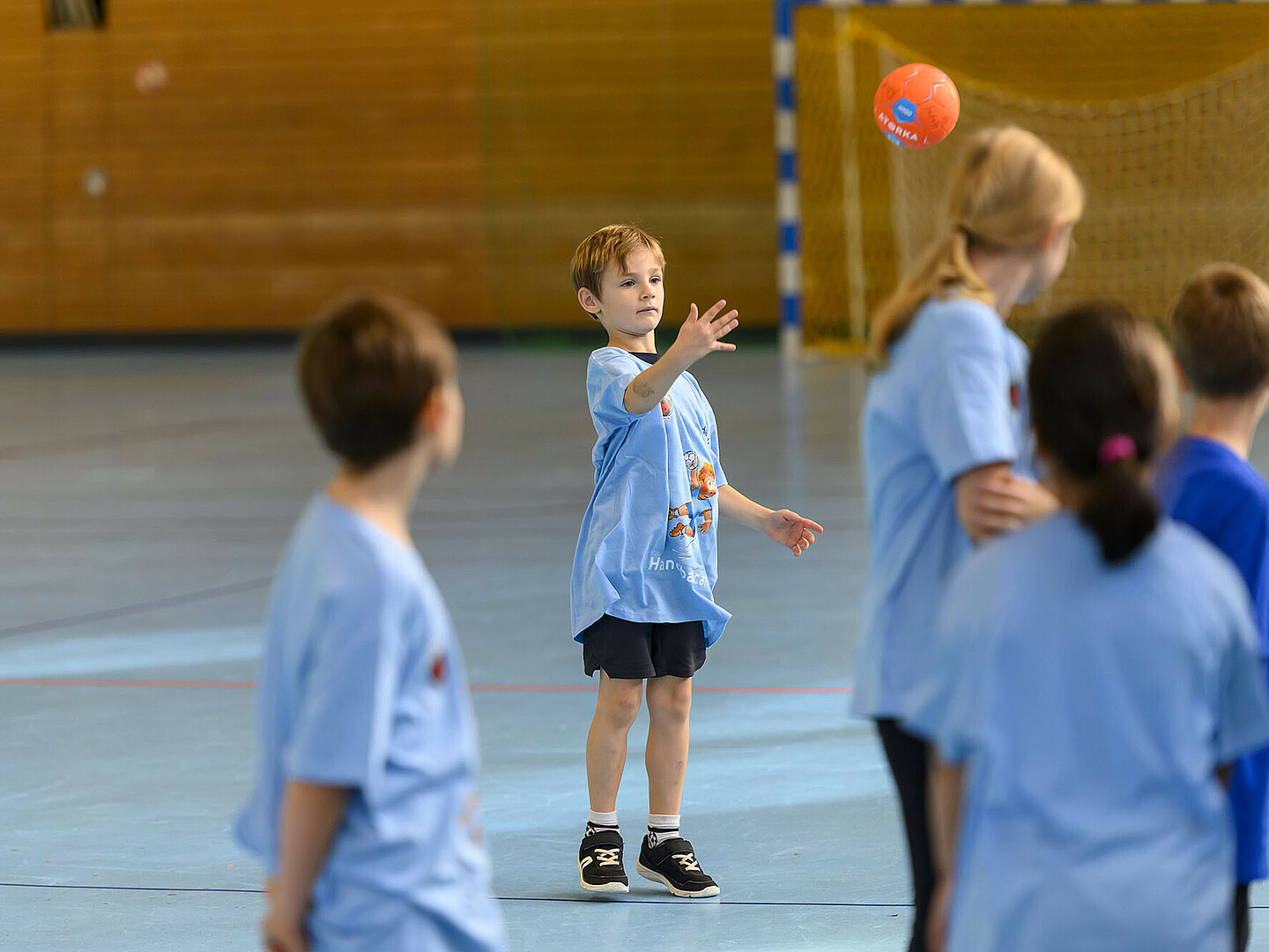 Kinder in blauen Trikots spielen in einer Sporthalle Handball.