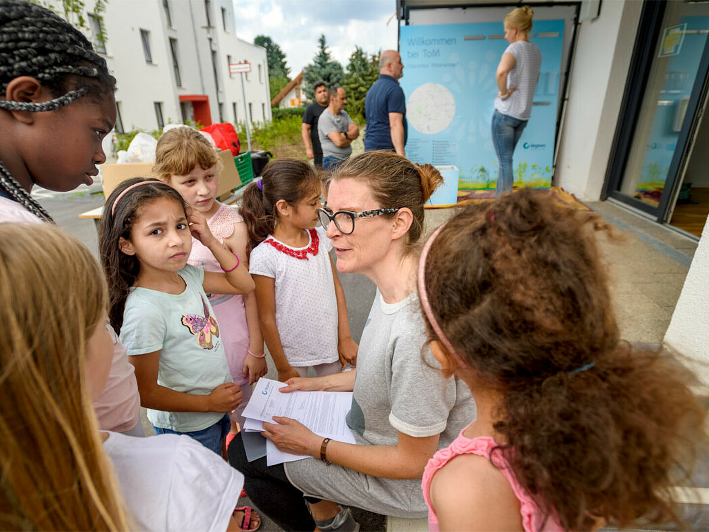 Eine Gruppe Kinder hat sich um eine erwachsene Frau versammelt, die ihnen etwas erklärt. Das Foto ist vor dem Wohnprojekt ToM – Tolerantes Miteinander aufgenommen, im Hintergrund stehen vier Erwachsene und unterhalten sich.