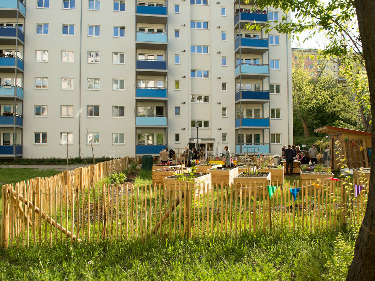 Vor einem mehrstöckigen Wohnhaus ist eine Grünfläche und eingezäunter Garten zu erkennen. In diesem halten sich Mieter des Hauses auf.