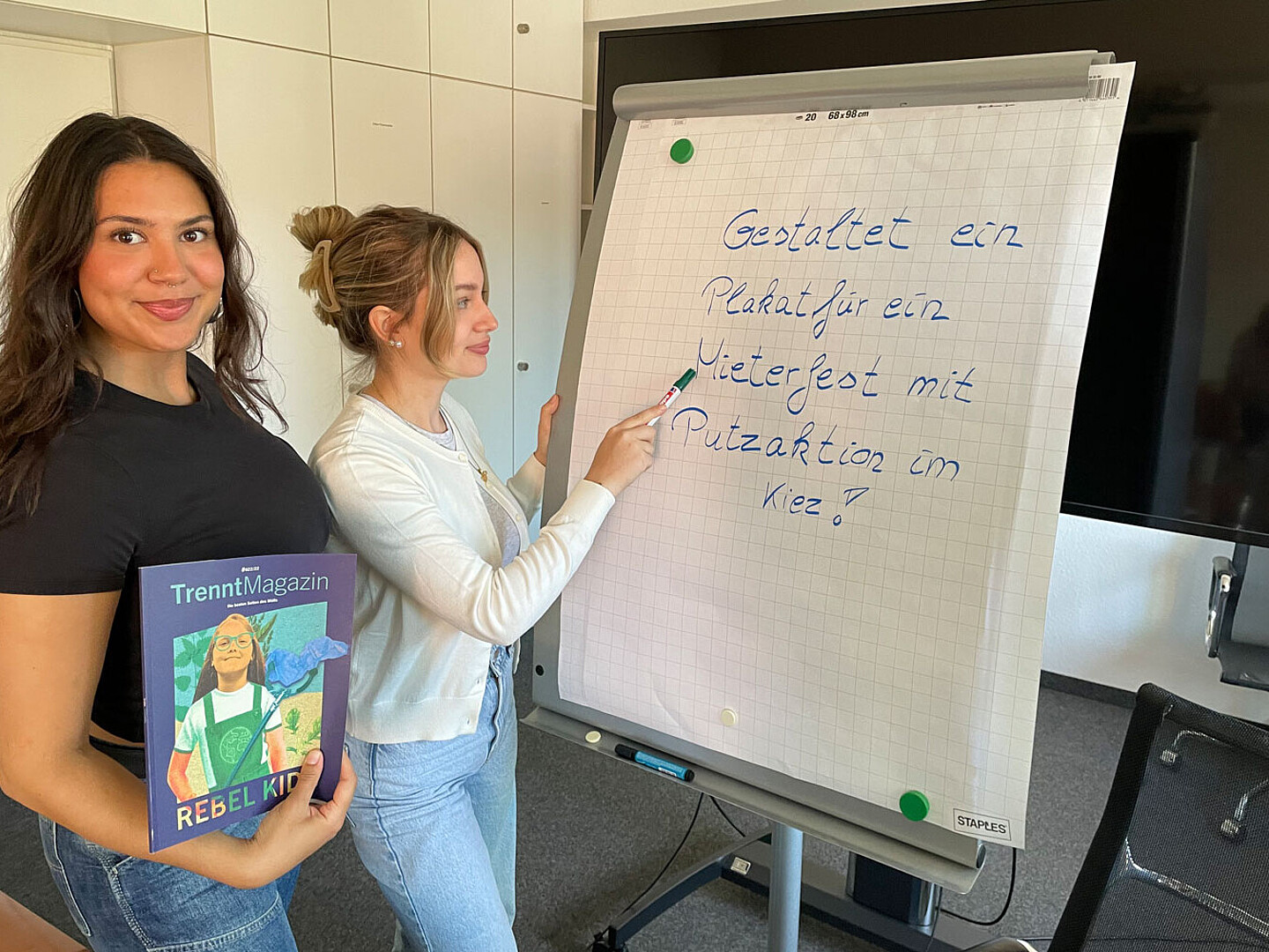 Zwei Auszubildende gestalten auf einem Flipchart ein Plakat. Eine Auszubildene hält ein Buch in die Kamera.