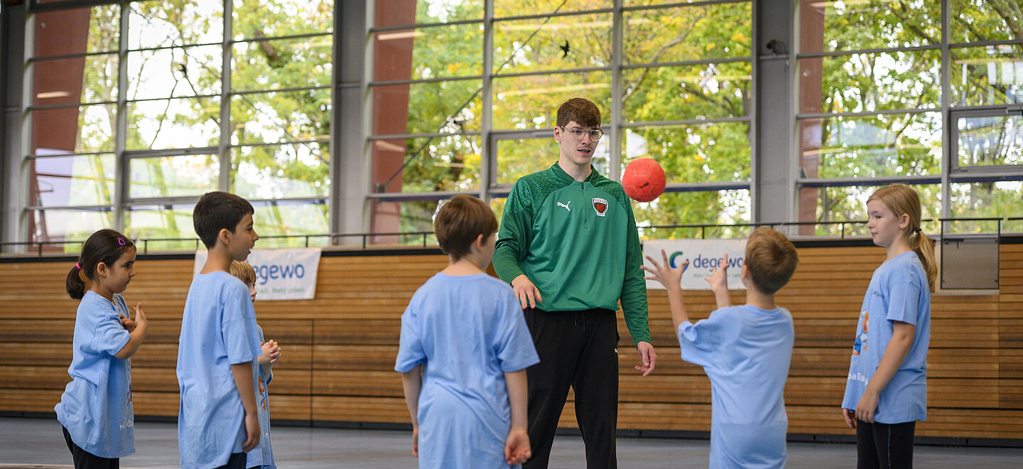 Eine Gruppe Kinder steht in einer Sporthalle mit ihrem Trainer. Ein Kind wirft einen kleinen roten Ball in Richtung Trainer.