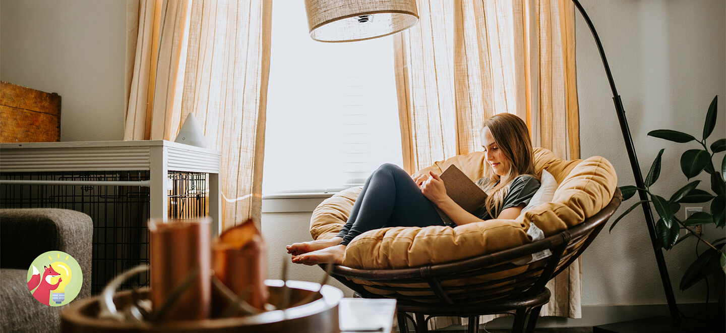 Eine Person sitzt zum Lesen in einem Sessel am Fenster. Das Zimmer ist mit warmen Farben dekoriert.