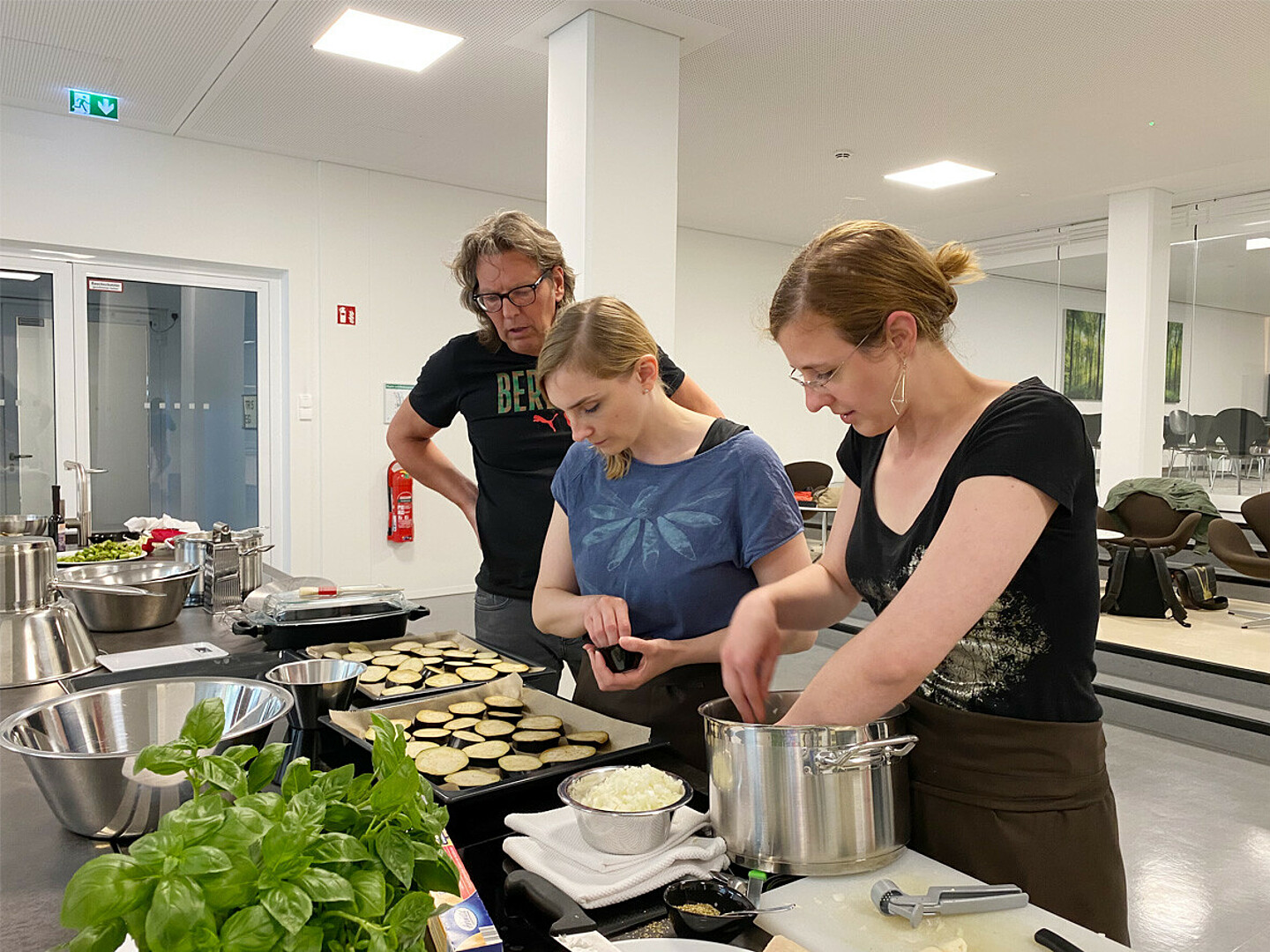 Zwei Frauen bestücken Backbleche mit Auberginenscheiben, ein Mann schaut zu.