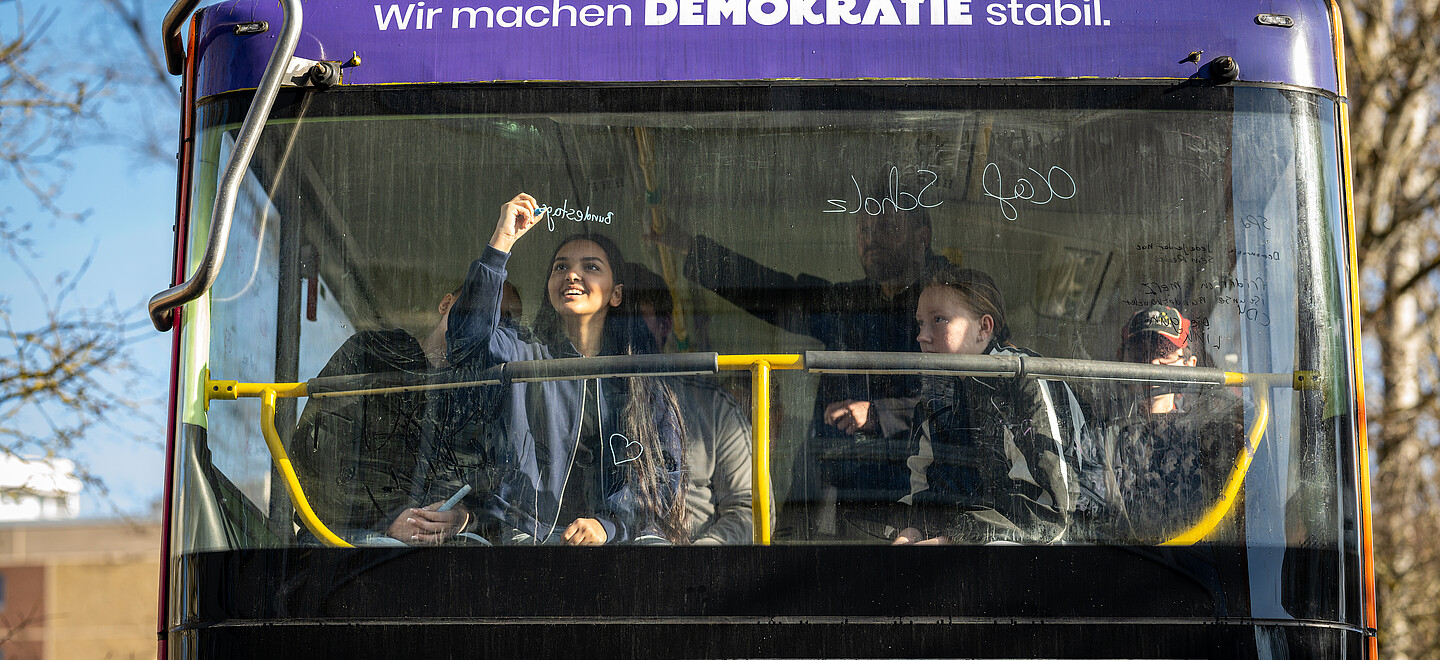 Blick von Außen auf die Frontscheibe eines Doppeldecker-Busses mit der Aufschrift: Wir machen Demokratie stabil. Hinter der Scheibe schreibt ein Mädchen etwas ans Fenster mit einem bunten Stift.