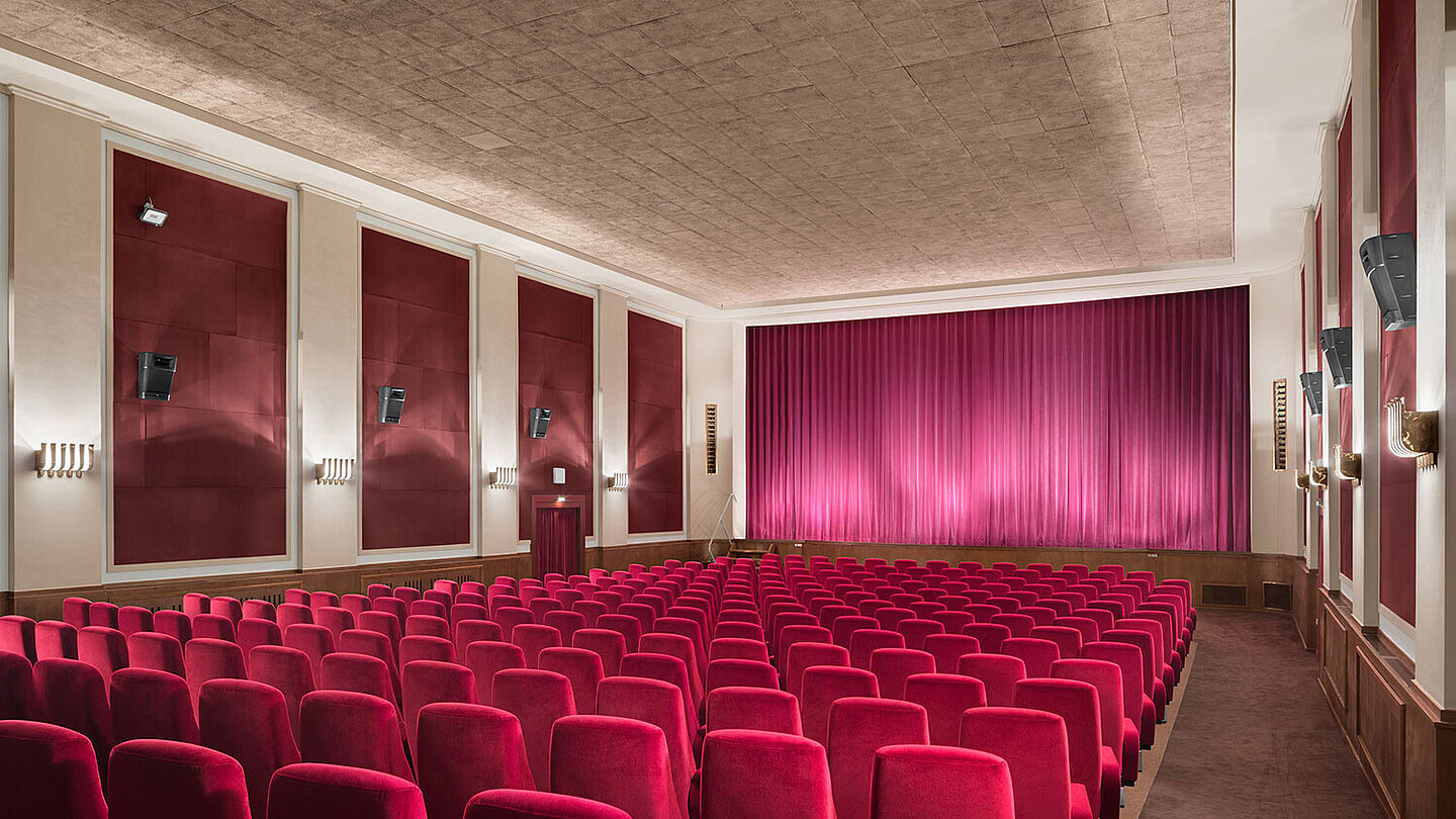 Blick in den Kinosaal des Odeon: Am Ende des Saals hängt ein großer pinker Vorhang vor der großen Leinwand. Davor Reihen roter Sessel. Der Boden und die rechteckigen Wandelemente zwischen kleinen Säulen greifen den dunklen Braunton der unteren Holzborte auf. Die Decke und restlichen Wandelemente sind cremefarben.