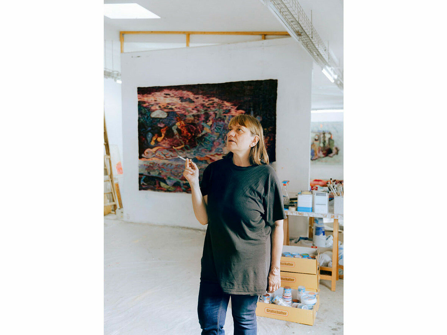 Die Künstlerin Kata Unger in ihrem großzügigen Atelier und ihre Arbeitskleidung. Sie hält einen Stift und zeigt auf die Wand