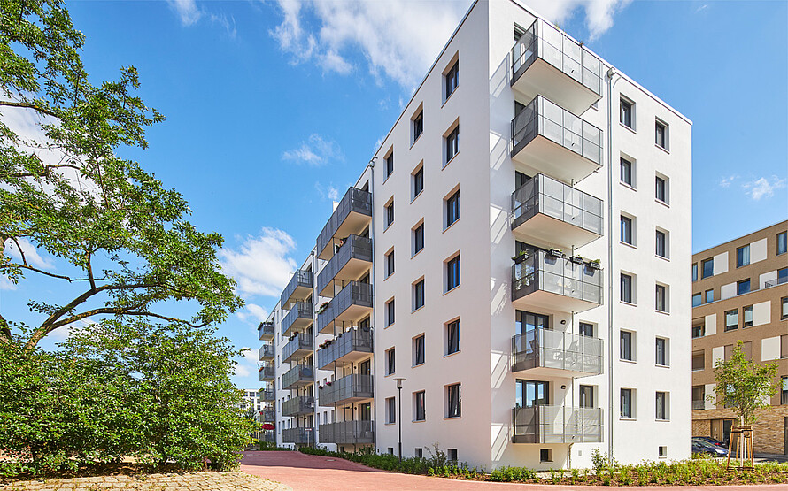 Blick auf ein Wohnhaus in der Sonne, die Balkone sind mit Blumen und Sonnenschirm gestaltet.