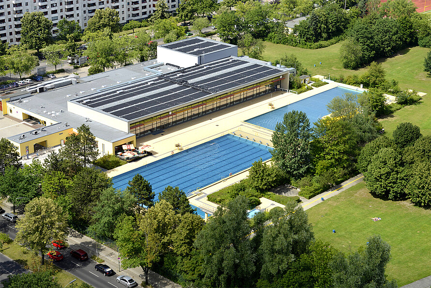 Luftaufnahme eines Schwimmbads, bestehend aus Hallen- und Freibad. Die zwei Becken im Außenbereich sind umringt von Bäumen und einer großen Grünfläche.