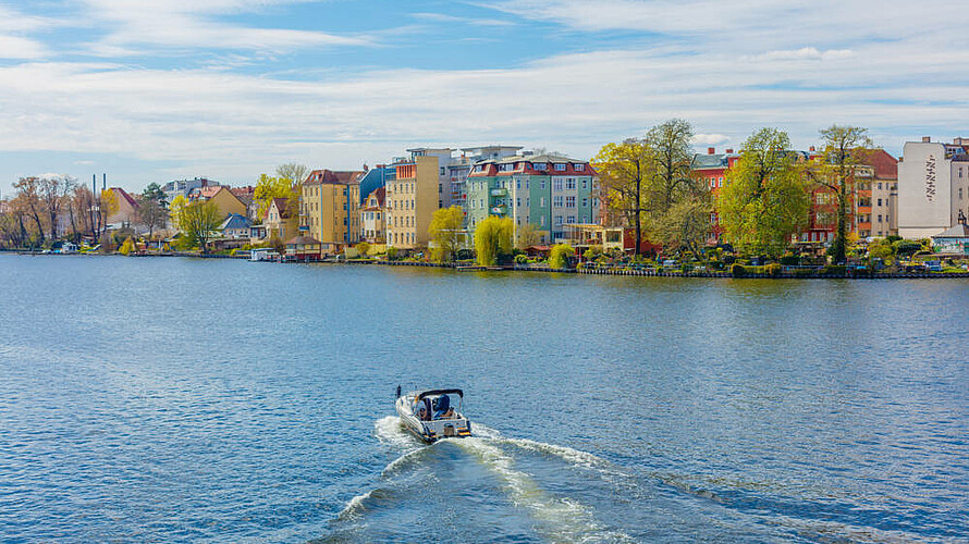 Blick auf einen Fluss bei sonnigem Wetter. Mittig fährt ein Motorboot. Am Ufer sind Altbauten und Bäume zu erkennen.