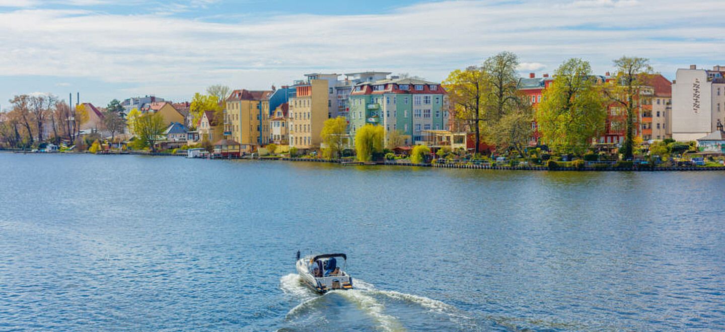 Blick auf einen Fluss bei sonnigem Wetter. Mittig fährt ein Motorboot. Am Ufer sind Altbauten und Bäume zu erkennen.