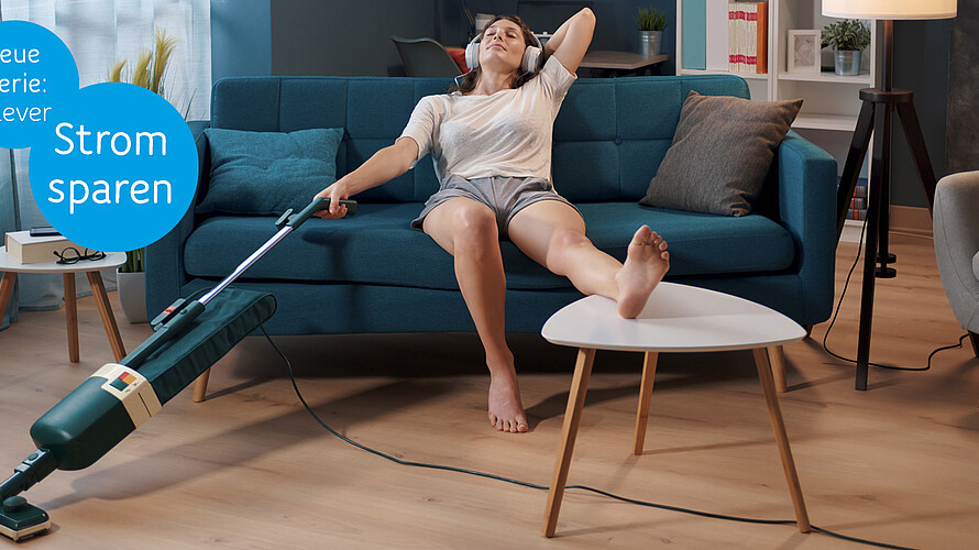 Eine Person sitzt im Wohnzimmer auf dem Sofa. Einen Fuß auf dem Couchtisch, hält sie in der rechten Hand einen Staubsauger und hört dabei über Kopfhörer Musik.