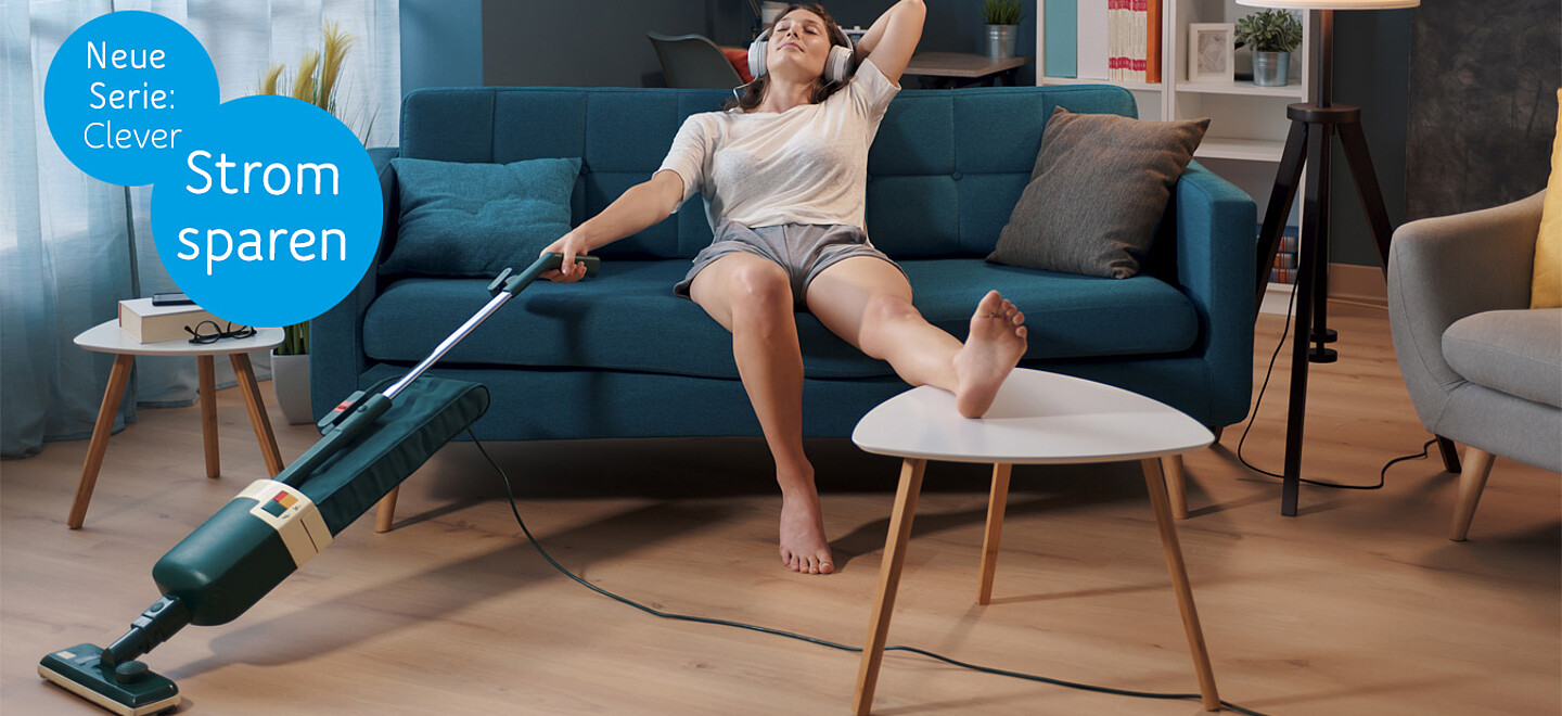 Eine Person sitzt im Wohnzimmer auf dem Sofa. Einen Fuß auf dem Couchtisch, hält sie in der rechten Hand einen Staubsauger und hört dabei über Kopfhörer Musik.