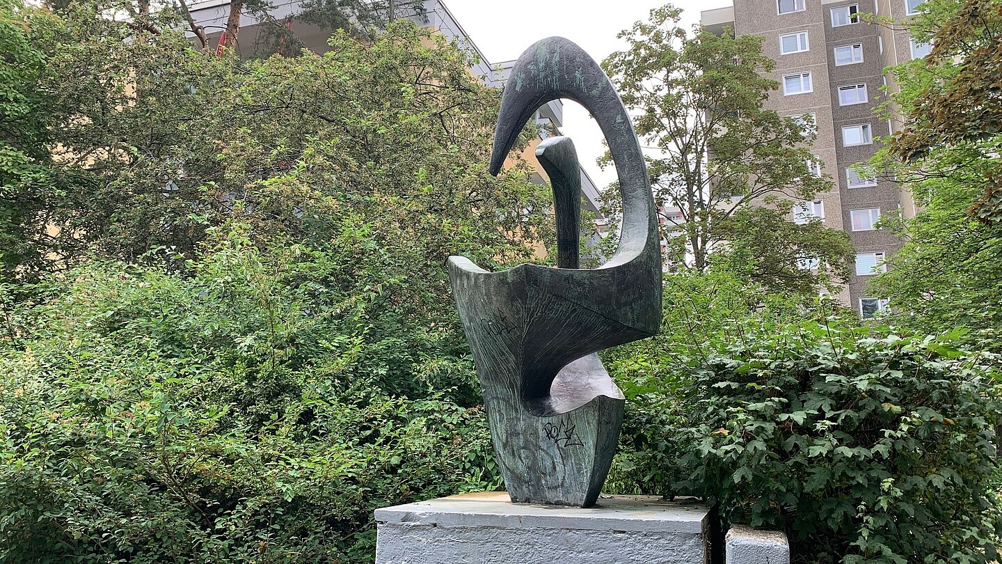 Eine abstrakte Skulptur aus oxidierter Bronze auf einem Betonsockel vor grünen Sträuchern. Im Hintergrund hohe Wohnhäuser.