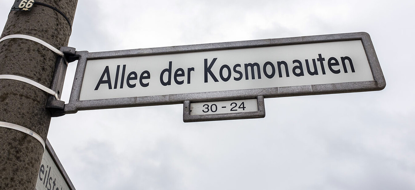 Strassenschild Alle der Kosmonauten: Schriften in schwarz vor dem weißen Hintergrund