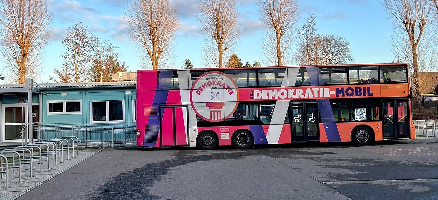 Bunt gestalteter Doppeldeckerbus "Demokratie-Mobil" steht vor Gebäuden und Bäumen.