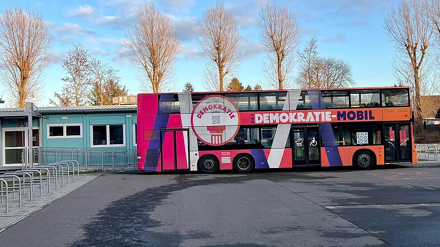 Bunt gestalteter Doppeldeckerbus "Demokratie-Mobil" steht vor Gebäuden und Bäumen.